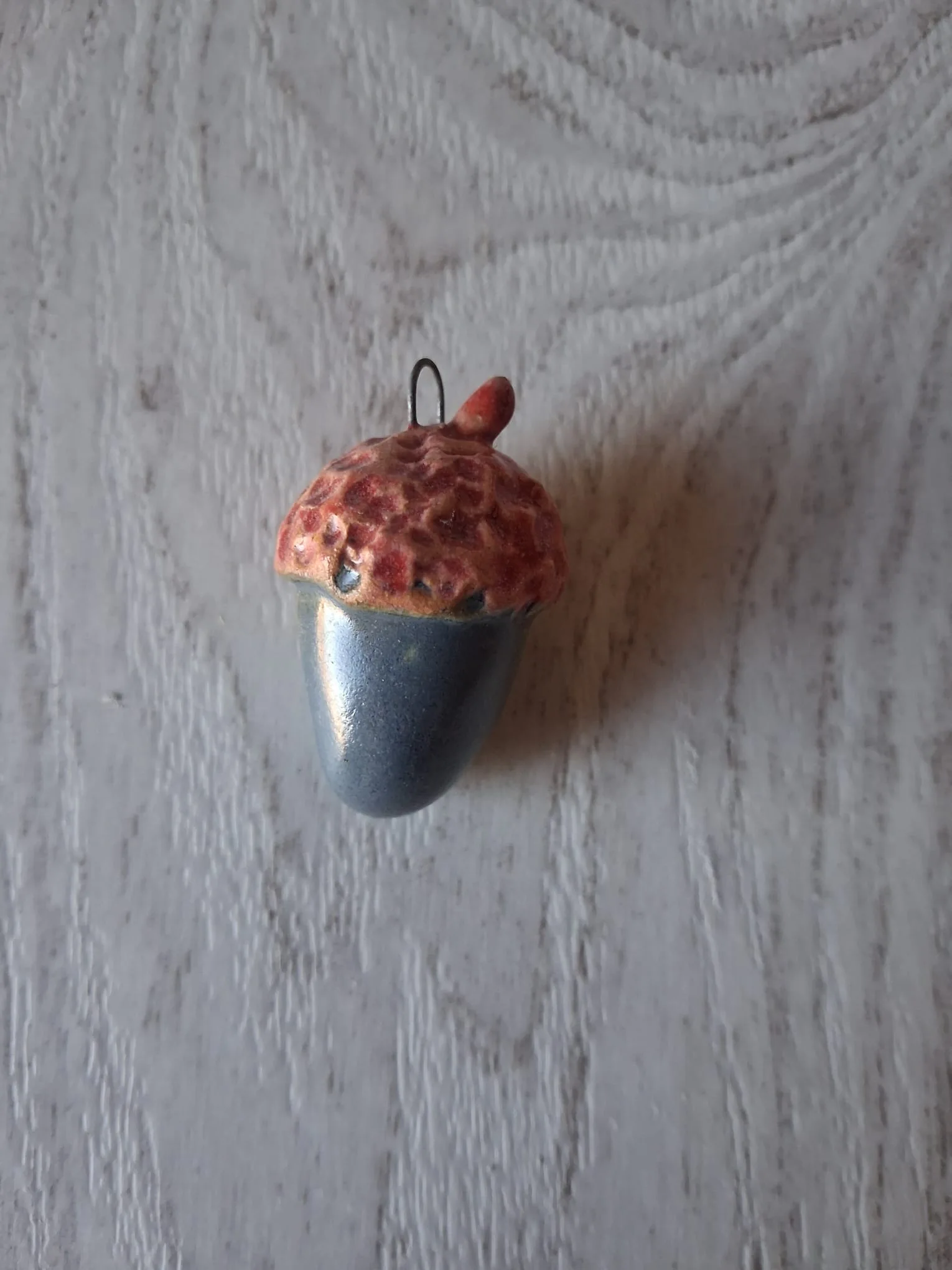 Adorno de Navidad de gres con forma de bellota de unos 4 cm, de cuerpo azul grisáceo brillante y capuchón rojo moteado, con anilla de alambre para colgar, esmaltado a mano en Valladolid — CreArte