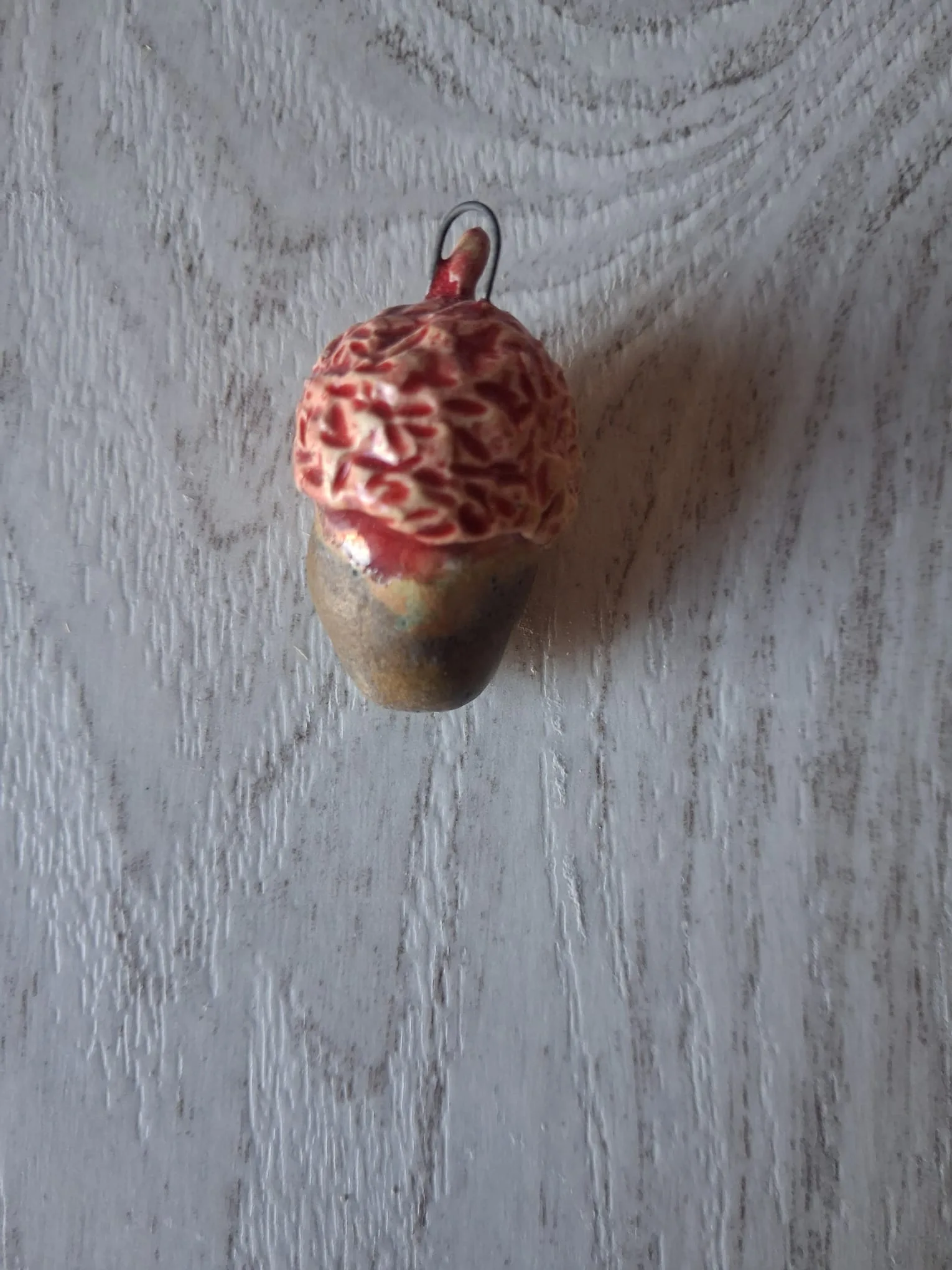 Adorno de Navidad de gres con forma de bellota de unos 4 cm, de capuchón rojo y crema con dibujo de hojas y cuerpo marrón verdoso, con anilla de alambre para colgar, esmaltado a mano en Valladolid — CreArte