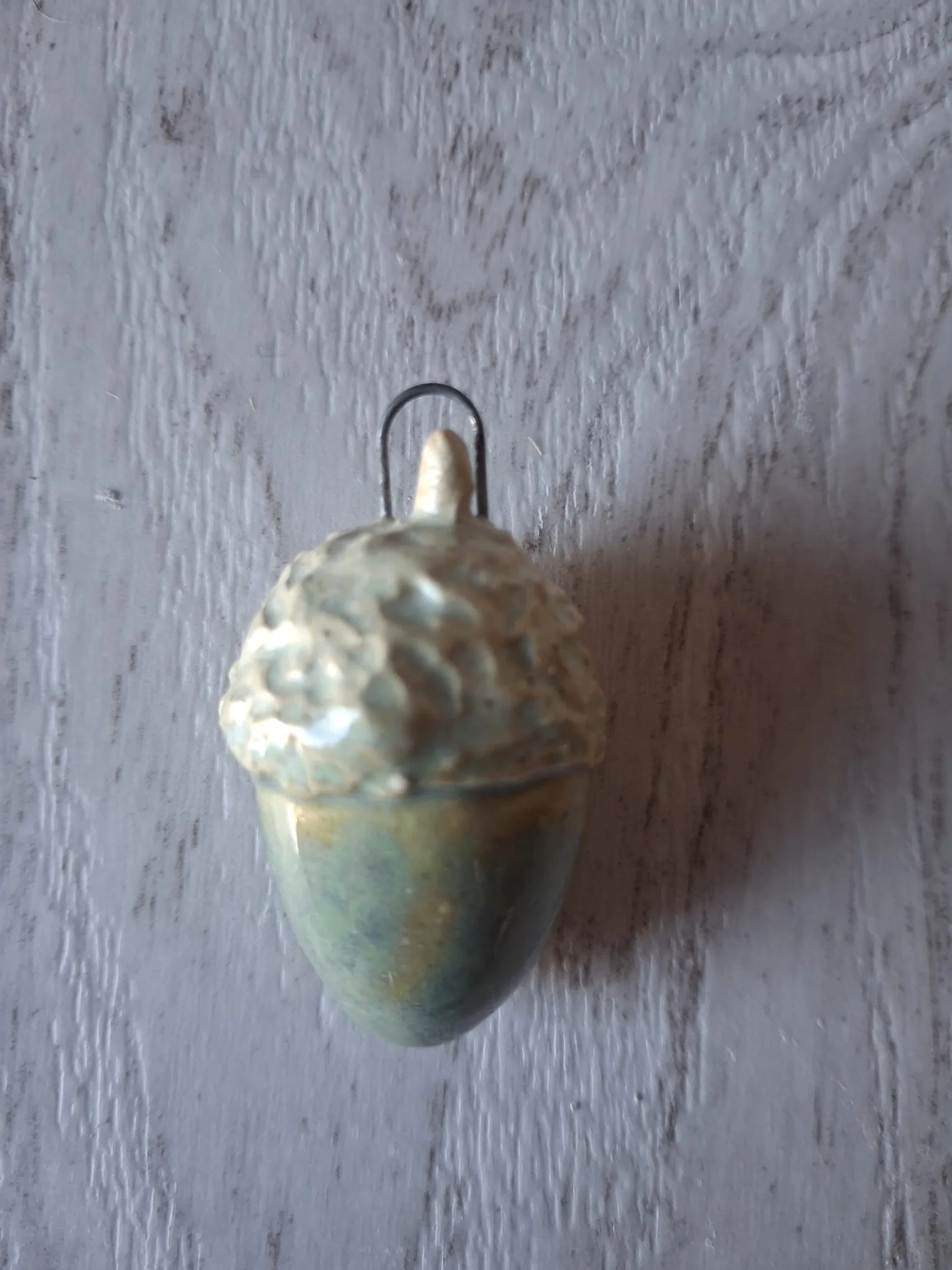 Adorno de Navidad de gres con forma de bellota de unos 4 cm, de cuerpo verde azulado con matices dorados y capuchón crema en relieve, con anilla de alambre para colgar, esmaltado a mano en Valladolid — CreArte