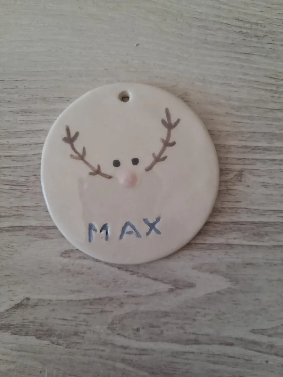 Adorno de Navidad de cerámica redondo con reno de cuernos marrones y nariz rosa y el nombre MAX pintado en azul, personalizado a mano en Valladolid — CreArte