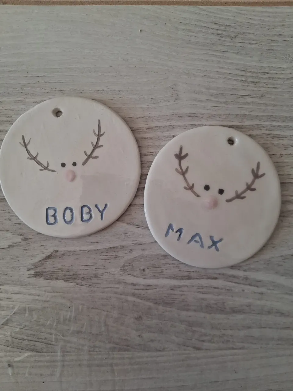 Dos adornos de Navidad de cerámica redondos con renos de cuernos marrones y nariz rosa y los nombres BOBY y MAX pintados en azul, personalizados a mano en Valladolid — CreArte