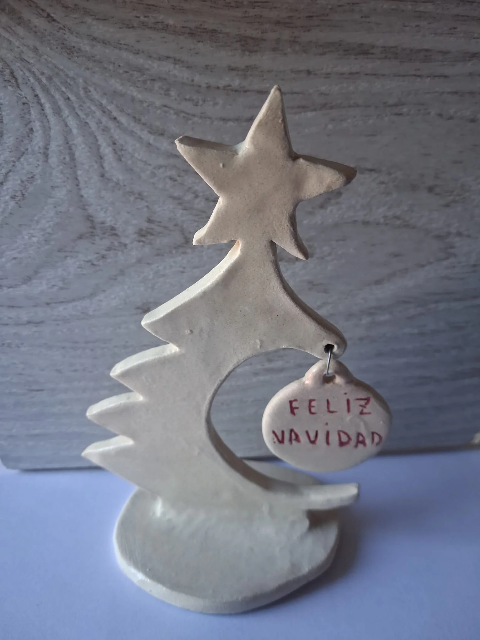 Árbol de Navidad de cerámica artesanal con estrella en la copa, base ovalada y un adorno colgante con el mensaje Feliz Navidad en rojo, hecho a mano en Valladolid — CreArte