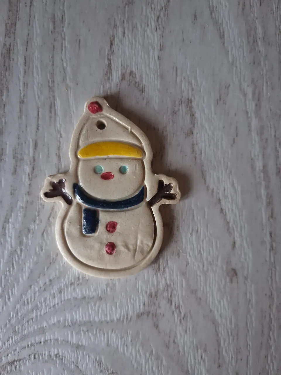 Figura de Navidad de cerámica con muñeco de nieve beige, gorro con pompón rojo y ribete amarillo, bufanda azul marino, brazos de ramita marrón y botones rojos, con agujero para colgar, de unos 5-6 cm, hecha a mano en Valladolid — CreArte