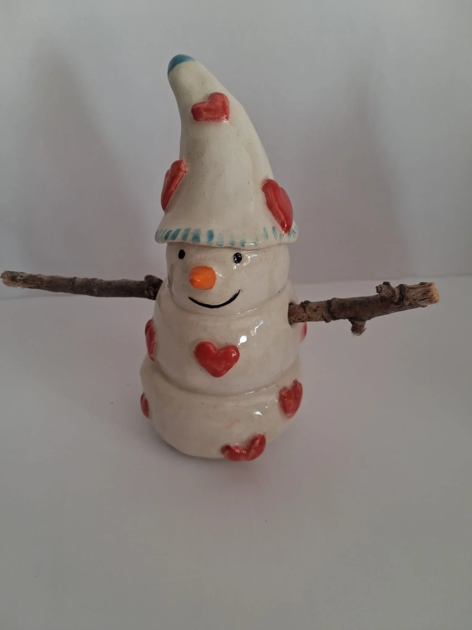 Muñeco de nieve de cerámica con gorro blanco de punta azul decorado con corazones rojos, nariz de zanahoria y brazos de ramitas, hecho a mano en Valladolid — CreArte