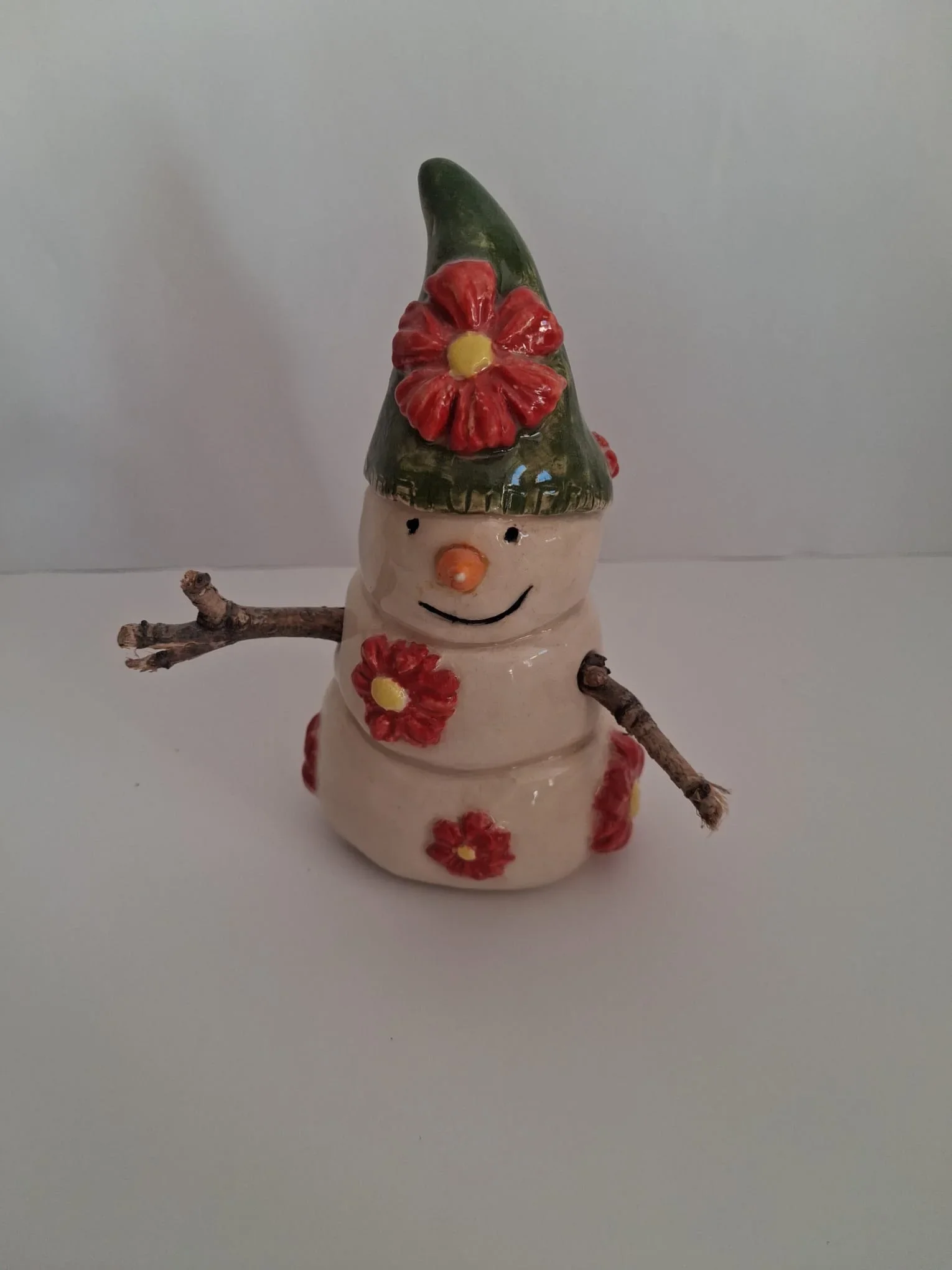 Muñeco de nieve de cerámica con gorro verde decorado con una flor roja, flores en el cuerpo, nariz naranja y brazos de ramitas, hecho a mano en Valladolid — CreArte