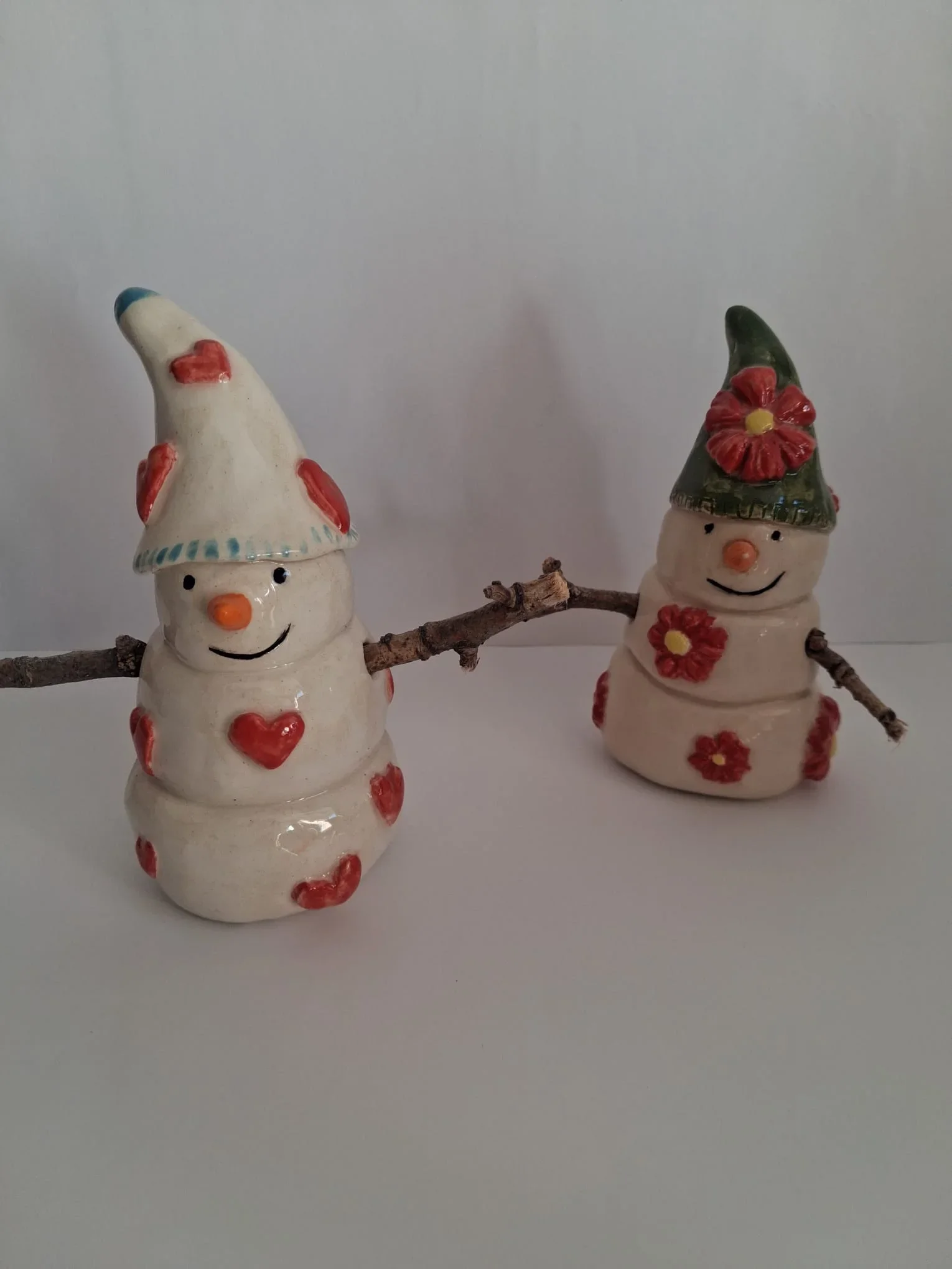 Dos muñecos de nieve de cerámica con brazos de ramitas: uno con gorro blanco y corazones rojos y otro con gorro verde y flores rojas, hechos a mano en Valladolid — CreArte