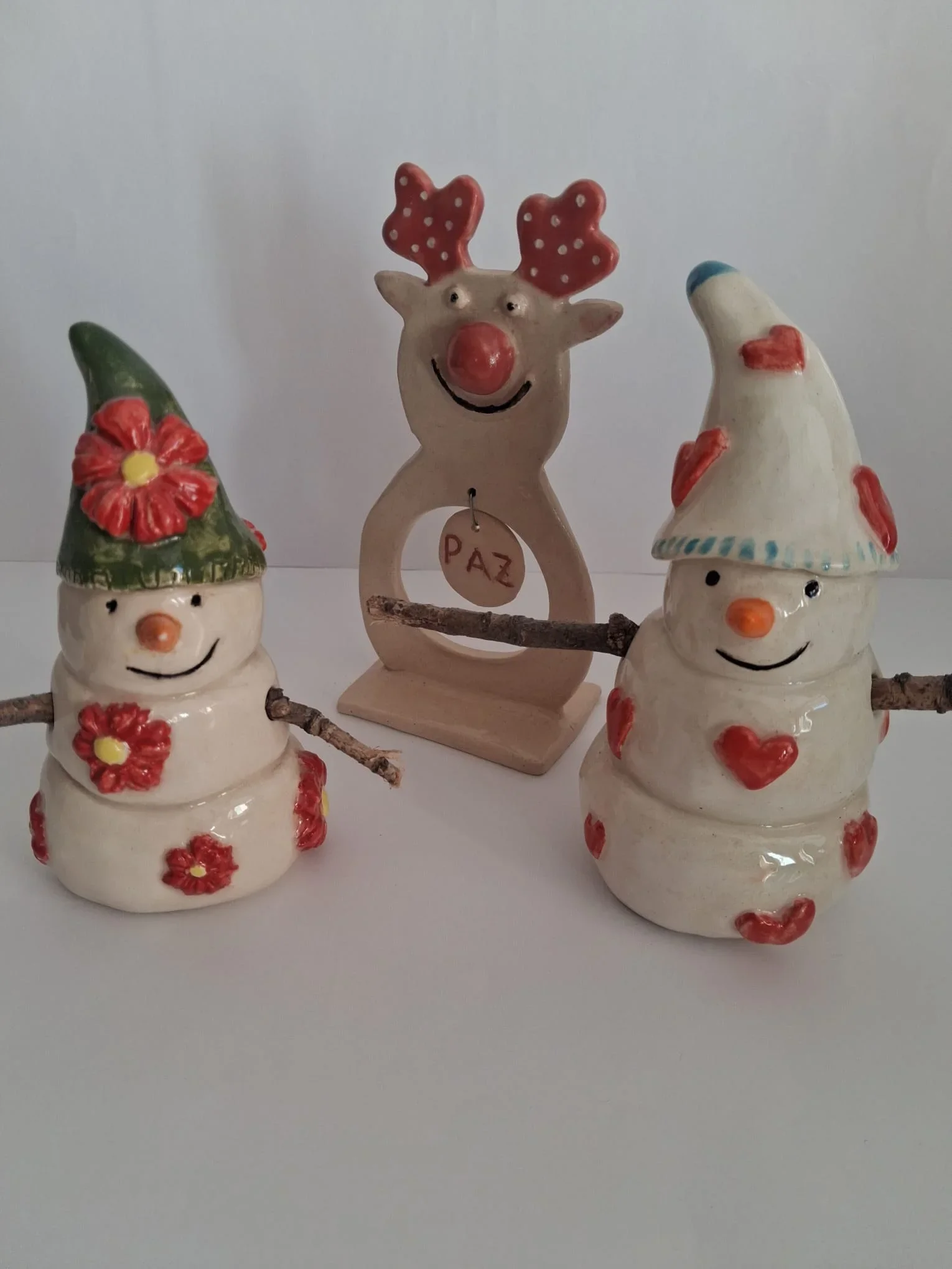 Reno de nariz roja con colgante PAZ junto a dos muñecos de nieve de cerámica, uno con gorro verde y flores y otro con gorro blanco y corazones, hechos a mano en Valladolid — CreArte