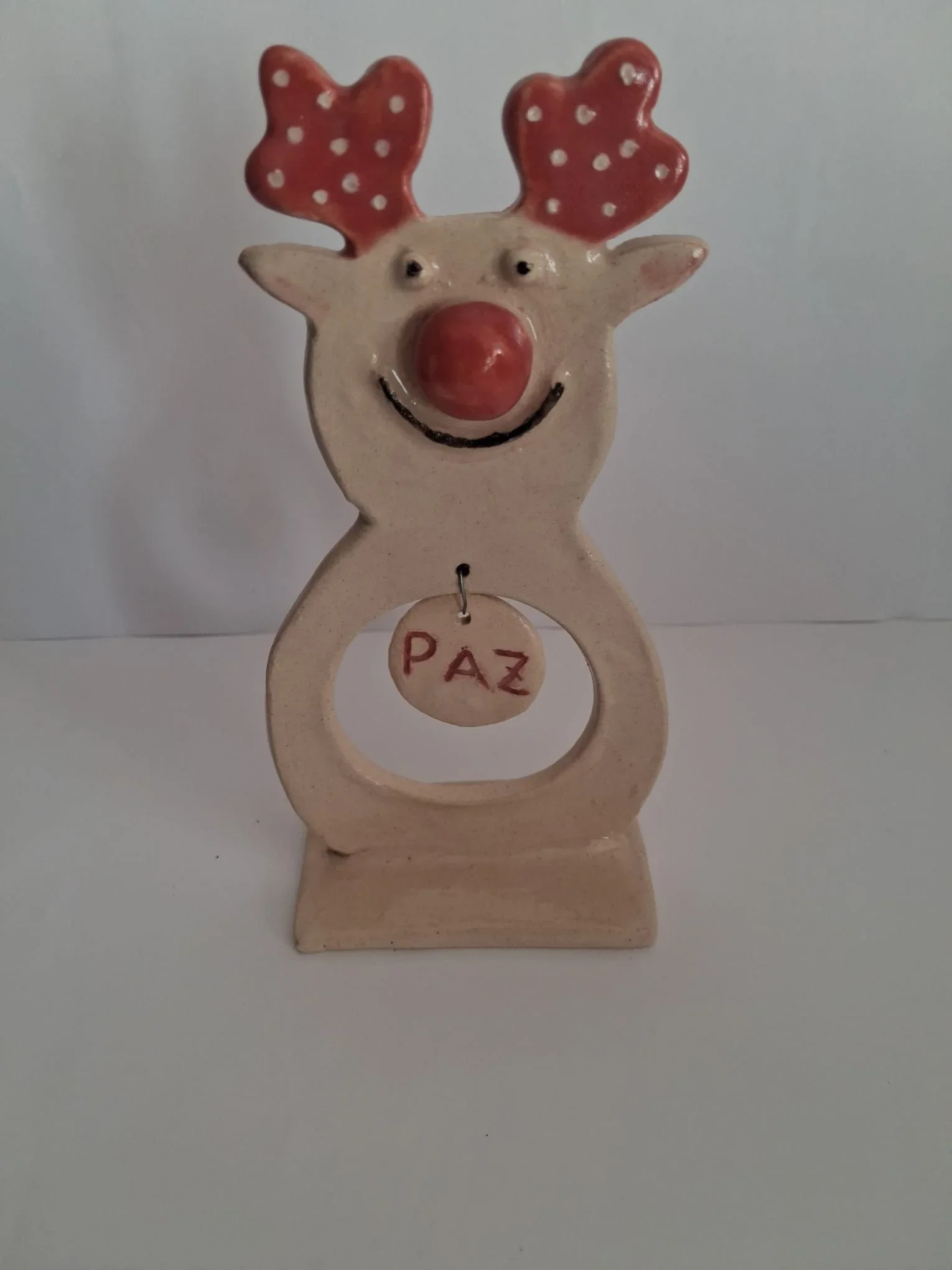 Figura de Navidad de cerámica con reno de nariz roja, astas rojas con lunares blancos y un colgante con la palabra PAZ, sobre base, vista frontal, hecha a mano en Valladolid — CreArte