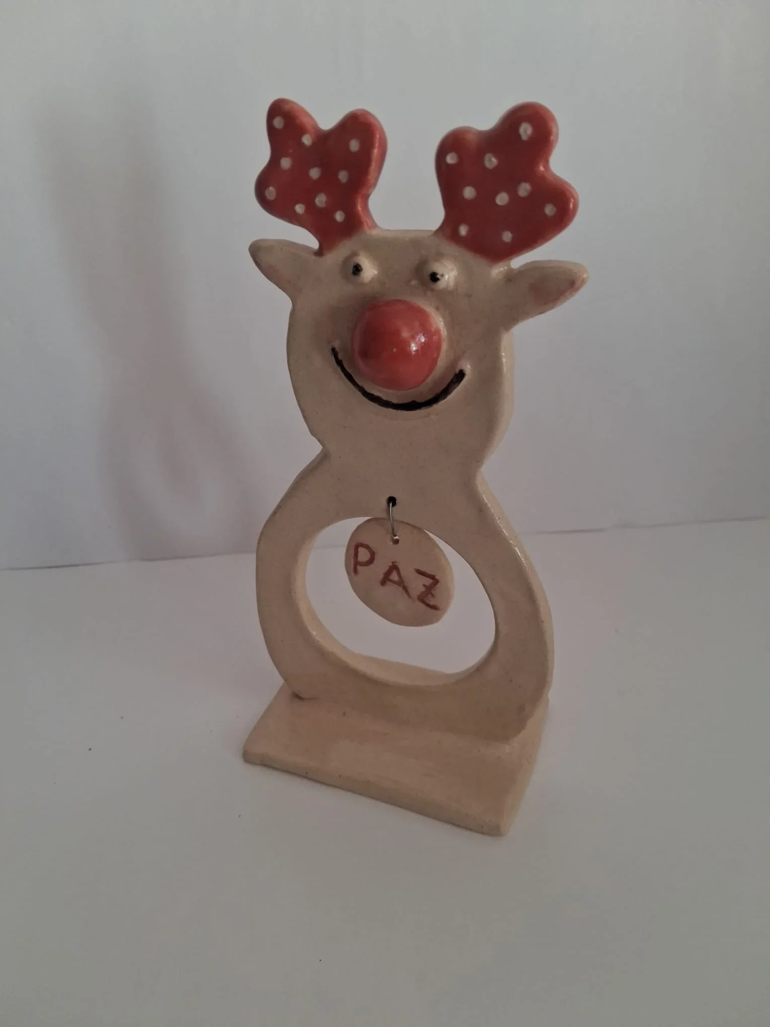 Figura de Navidad de cerámica con reno de nariz roja, astas con lunares y colgante PAZ sobre base, vista lateral, hecha a mano en Valladolid — CreArte